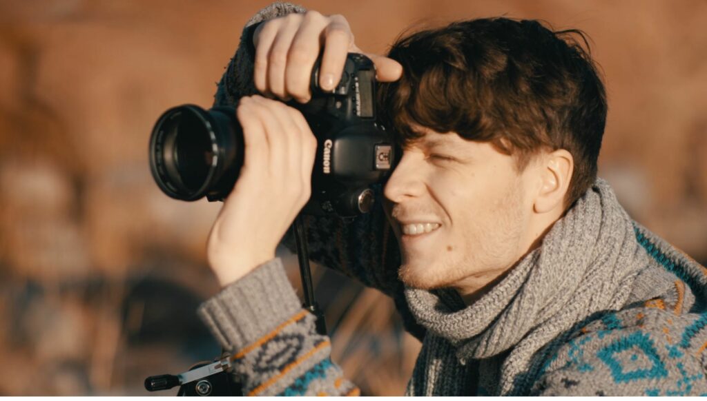 Lähikuva hymyilevästä villapaitapukeisesta miehestä joka on ottamassa Canonin järjestelmäkameralla valokuvaa.