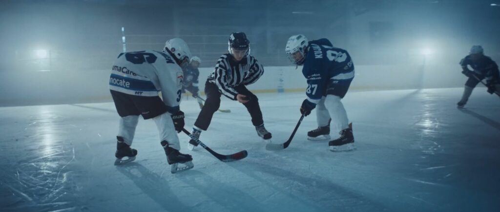 Jääkiekkotuomari tiputtamassa kiekkoa jäälle, kaksi pelaajaa seisoo valmiudessa mailat kädessä.