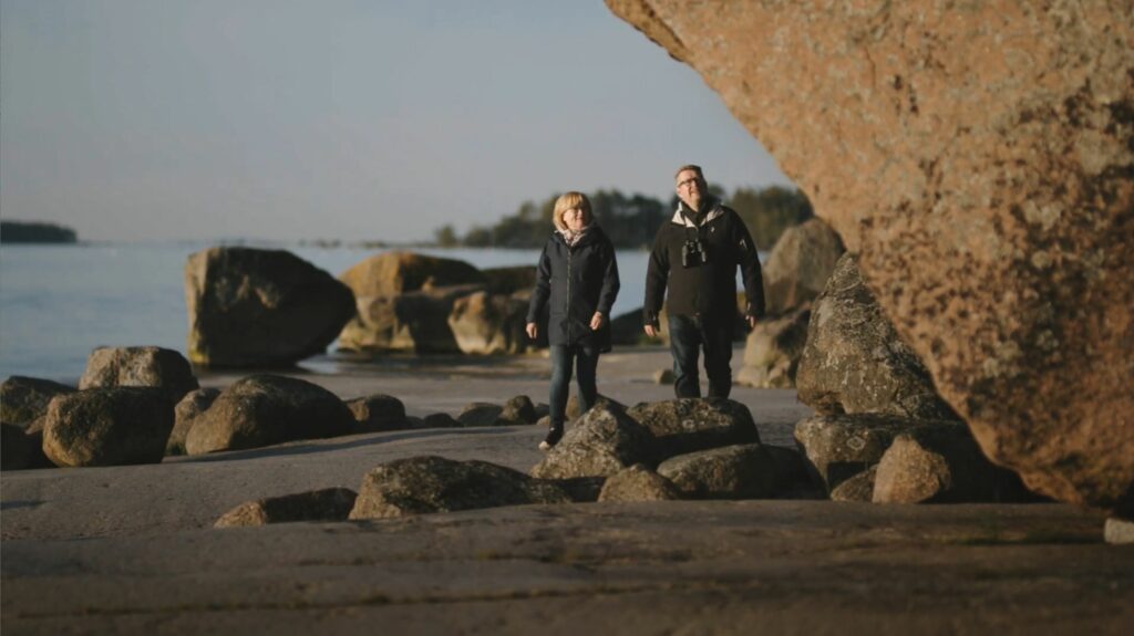 Nainen ja mies kävelee ulkoiluvaatteissa rannalla ison kiven vieressä. Paljon pieniä kiviä taustalla.