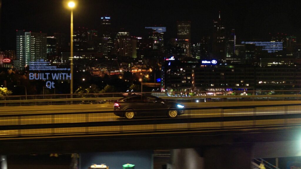 Auto ajaa yksin monikaistaisella tiellä illalla. Taustalla suurkaupunki.