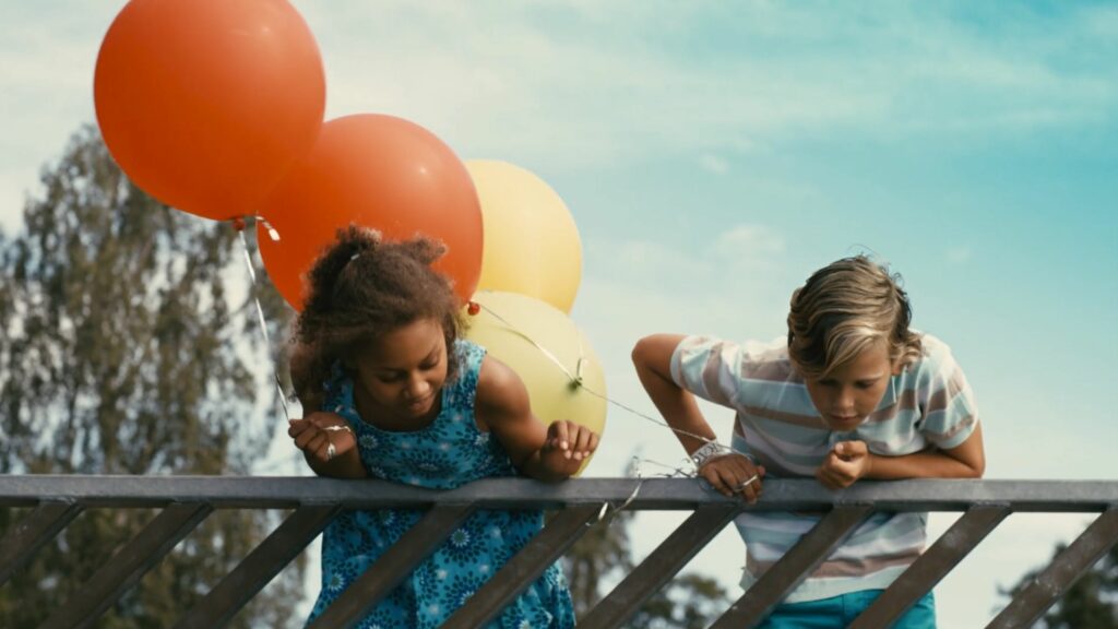 Kaksi lasta kesäpäivänä nojaa kaiteen yli ja katsoo alaspäin. Molemmilla kädessään ilmapalloja.