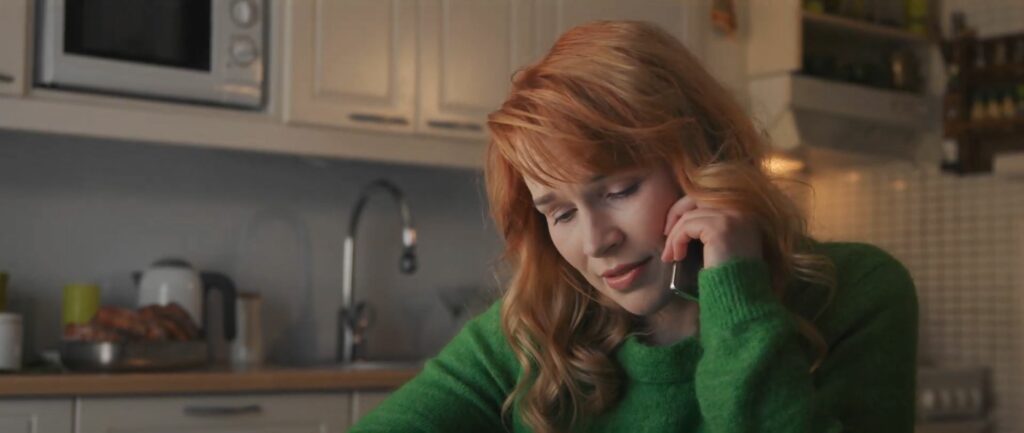 Lähikuva punatukkaisesta naisesta, joka katsoo alaviistoon puhuessaan puhelimessa keittiössä.