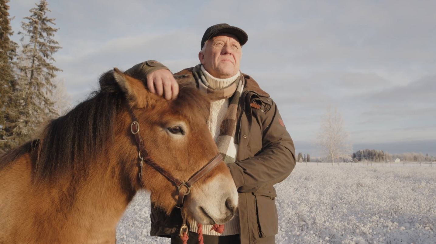 Vanha mies seisoo talvisessa maisemassa ja rapsuttaa hevosen otsaa.